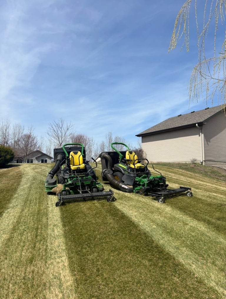 Two yellow and green riding lawn mowers on a striped mowed lawn near a white residential building
