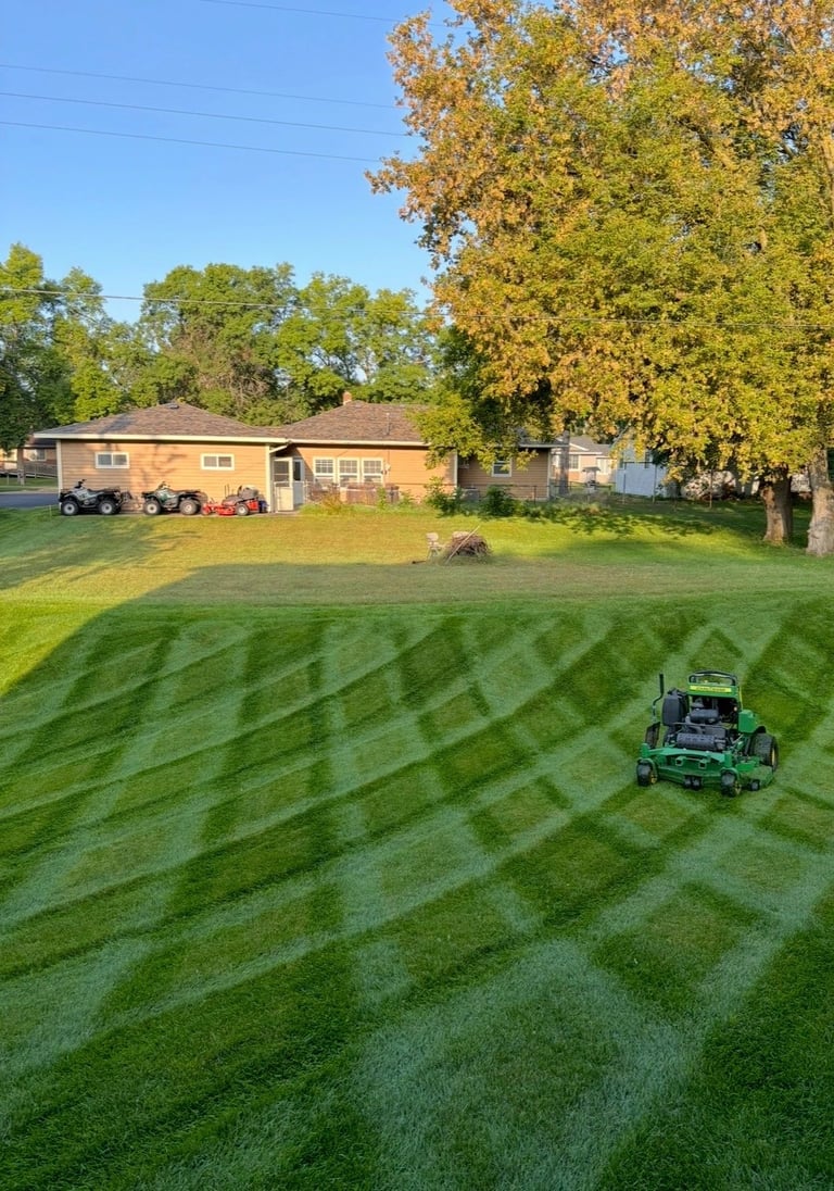 Green striped lawn being mowed by a riding mower in front of a brick house with trees